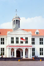 A classic colonial-style building with a central tower and a red tile roof. The building has large windows and a prominent entrance with columns. The Indonesian flag is displayed in front, and several people are visible near the structure. There are clear skies and some small trees or shrubs are positioned near the building.