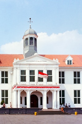 A classic colonial-style building with a central tower and a red tile roof. The building has large windows and a prominent entrance with columns. The Indonesian flag is displayed in front, and several people are visible near the structure. There are clear skies and some small trees or shrubs are positioned near the building.