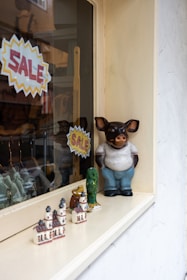 A shop window display featuring ceramic figurines, including a large anthropomorphic pig wearing a shirt and pants. Smaller decorative items, such as miniature buildings and animal figures, are also present. Two bright yellow and red 'SALE' signs are visible.
