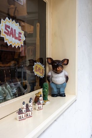 A shop window display featuring ceramic figurines, including a large anthropomorphic pig wearing a shirt and pants. Smaller decorative items, such as miniature buildings and animal figures, are also present. Two bright yellow and red 'SALE' signs are visible.