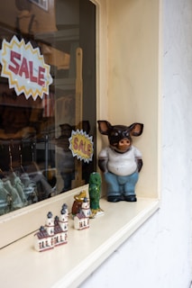 A shop window display featuring ceramic figurines, including a large anthropomorphic pig wearing a shirt and pants. Smaller decorative items, such as miniature buildings and animal figures, are also present. Two bright yellow and red 'SALE' signs are visible.