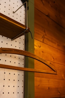 A wooden clothes hanger with the text 'AMERICAN SANITARY LAUNDRY' is hanging from a metal hook. The background features a wooden panel on the right and a perforated white wall on the left.