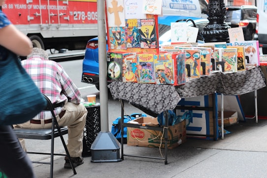 A street vendor stall displaying a variety of comic books on a table covered with a patterned cloth. The comics feature vibrant and colorful covers, with titles visible such as Wolverine and Arak. A person in a plaid shirt is seated on a chair beside the stall, with their back to the camera. In the background, a red delivery truck and a blue car are parked on the street.