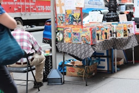 A street vendor stall displaying a variety of comic books on a table covered with a patterned cloth. The comics feature vibrant and colorful covers, with titles visible such as Wolverine and Arak. A person in a plaid shirt is seated on a chair beside the stall, with their back to the camera. In the background, a red delivery truck and a blue car are parked on the street.