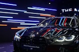 A sleek, black BMW car with reflective surfaces showcasing colorful LED strip lighting in the background. The lights are predominantly blue, red, and white. The car's headlights are on, displaying a modern and aggressive design, while a banner at the top reads 'BMW M Performance Parts.'