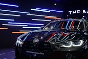 A sleek, black BMW car with reflective surfaces showcasing colorful LED strip lighting in the background. The lights are predominantly blue, red, and white. The car's headlights are on, displaying a modern and aggressive design, while a banner at the top reads 'BMW M Performance Parts.'