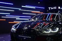 A sleek, black BMW car with reflective surfaces showcasing colorful LED strip lighting in the background. The lights are predominantly blue, red, and white. The car's headlights are on, displaying a modern and aggressive design, while a banner at the top reads 'BMW M Performance Parts.'