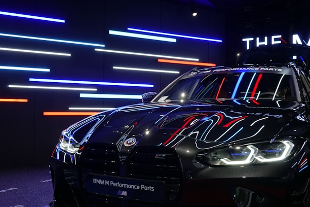 A sleek, black BMW car with reflective surfaces showcasing colorful LED strip lighting in the background. The lights are predominantly blue, red, and white. The car's headlights are on, displaying a modern and aggressive design, while a banner at the top reads 'BMW M Performance Parts.'