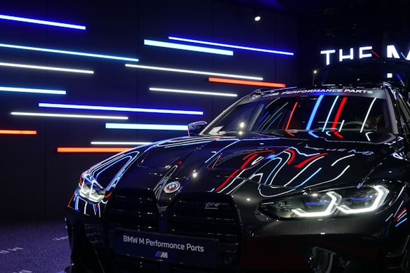 A sleek, black BMW car with reflective surfaces showcasing colorful LED strip lighting in the background. The lights are predominantly blue, red, and white. The car's headlights are on, displaying a modern and aggressive design, while a banner at the top reads 'BMW M Performance Parts.'