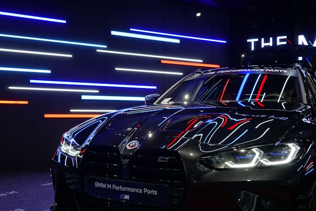 A sleek, black BMW car with reflective surfaces showcasing colorful LED strip lighting in the background. The lights are predominantly blue, red, and white. The car's headlights are on, displaying a modern and aggressive design, while a banner at the top reads 'BMW M Performance Parts.'