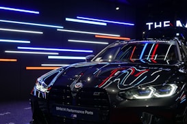 A sleek, black BMW car with reflective surfaces showcasing colorful LED strip lighting in the background. The lights are predominantly blue, red, and white. The car's headlights are on, displaying a modern and aggressive design, while a banner at the top reads 'BMW M Performance Parts.'