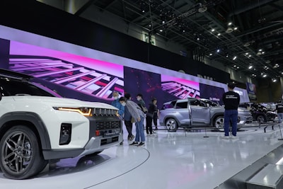 A car exhibition displays several modern vehicles on a brightly lit showroom floor. People are inspecting the cars, which feature a sleek, futuristic design with prominent grilles and stylish body work. A large digital screen in the background shows vibrant visuals that add to the high-tech atmosphere of the event.