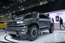 A sleek, modern SUV is displayed prominently on a showroom floor. It features a matte grey finish with bold, angular lines and rugged tires, indicative of off-road capability. Bright lights illuminate the vehicle, highlighting its design elements. In the background, people are walking by and there is a large digital screen displaying an advertisement.