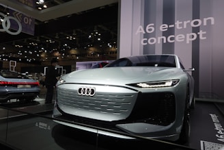 A sleek, futuristic car is prominently displayed under bright lights in an exhibition space, with a large backdrop showcasing the words 'A6 e-tron concept.' The car features a shiny metallic finish, distinctive LED headlights, and a minimalist, aerodynamic design. Multiple reflections are visible on the car's surface, indicating the strong ambient lighting.