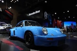 A classic blue Porsche is displayed in an indoor exhibition setting with prominent illuminations. The environment is a car show with Mercedes-Benz and Porsche branding visible in the backdrop. Other vehicles are present, adding to the auto show ambiance.