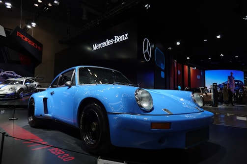 A classic blue Porsche is displayed in an indoor exhibition setting with prominent illuminations. The environment is a car show with Mercedes-Benz and Porsche branding visible in the backdrop. Other vehicles are present, adding to the auto show ambiance.