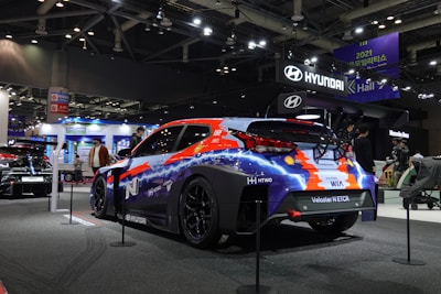 A vibrant, customized Hyundai race car displayed at a car exhibition, featuring a colorful livery of blue, orange, and white. The setting is a well-lit showroom with other automobiles and signs, including prominent Hyundai branding. People are casually walking around, indicating a public event.