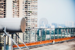 A rooftop view featuring a large cylindrical industrial vent or exhaust system in the foreground, with a glass railing along the edge. In the background, there are high-rise apartment buildings and an iconic circular building, indicative of an urban cityscape.