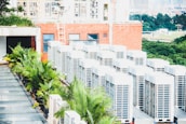 A row of air conditioning units is situated on a rooftop next to a red brick building. Several green palm plants are lined up along the side, providing a contrast to the industrial appearance of the AC units. In the background, a landscape of trees and distant buildings is visible, suggesting an urban setting.