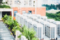 A row of air conditioning units is situated on a rooftop next to a red brick building. Several green palm plants are lined up along the side, providing a contrast to the industrial appearance of the AC units. In the background, a landscape of trees and distant buildings is visible, suggesting an urban setting.