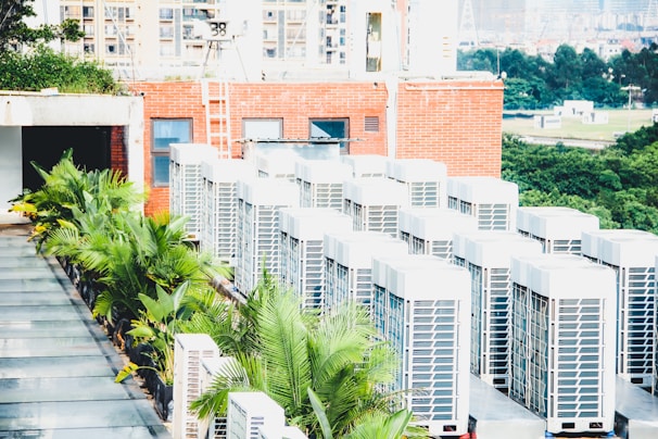 A row of air conditioning units is situated on a rooftop next to a red brick building. Several green palm plants are lined up along the side, providing a contrast to the industrial appearance of the AC units. In the background, a landscape of trees and distant buildings is visible, suggesting an urban setting.