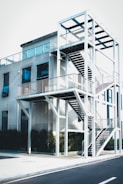 A modern building with an external staircase structure made of white metal, featuring multiple levels and railing. The building's exterior has large glass windows and the structure is surrounded by greenery at the base.