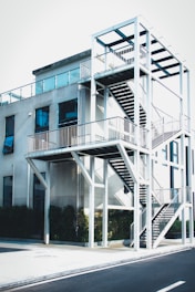 A modern building with an external staircase structure made of white metal, featuring multiple levels and railing. The building's exterior has large glass windows and the structure is surrounded by greenery at the base.