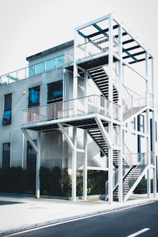 A modern building with an external staircase structure made of white metal, featuring multiple levels and railing. The building's exterior has large glass windows and the structure is surrounded by greenery at the base.