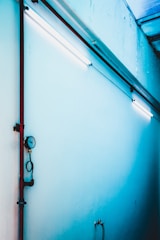 A brightly lit blue wall features a red vertical pipe with an attached gauge, alongside fluorescent lights affixed to the ceiling. The setting appears industrial with stark lines and minimalistic design elements.