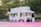 A colorful food stand offers a variety of treats such as Sno Kones, popcorn, caramel apples, funnel cakes, and cotton candy. The stand is adorned with vibrant signage and is parked on a pink surface surrounded by trees and a wooden fence. It has a classic, nostalgic design, reminiscent of fairground or carnival booths.