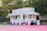 A colorful food stand offers a variety of treats such as Sno Kones, popcorn, caramel apples, funnel cakes, and cotton candy. The stand is adorned with vibrant signage and is parked on a pink surface surrounded by trees and a wooden fence. It has a classic, nostalgic design, reminiscent of fairground or carnival booths.