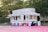 A colorful food stand offers a variety of treats such as Sno Kones, popcorn, caramel apples, funnel cakes, and cotton candy. The stand is adorned with vibrant signage and is parked on a pink surface surrounded by trees and a wooden fence. It has a classic, nostalgic design, reminiscent of fairground or carnival booths.