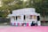 A colorful food stand offers a variety of treats such as Sno Kones, popcorn, caramel apples, funnel cakes, and cotton candy. The stand is adorned with vibrant signage and is parked on a pink surface surrounded by trees and a wooden fence. It has a classic, nostalgic design, reminiscent of fairground or carnival booths.