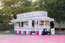 A colorful food stand offers a variety of treats such as Sno Kones, popcorn, caramel apples, funnel cakes, and cotton candy. The stand is adorned with vibrant signage and is parked on a pink surface surrounded by trees and a wooden fence. It has a classic, nostalgic design, reminiscent of fairground or carnival booths.