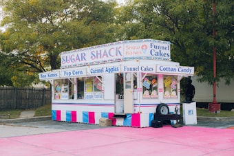 A colorful food stand offers a variety of treats such as Sno Kones, popcorn, caramel apples, funnel cakes, and cotton candy. The stand is adorned with vibrant signage and is parked on a pink surface surrounded by trees and a wooden fence. It has a classic, nostalgic design, reminiscent of fairground or carnival booths.