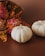 Two small white pumpkins are placed on a brown surface next to a bouquet of vibrant flowers. The flowers include pink and orange blossoms, accented with small orange filler flowers, creating an autumnal arrangement.