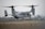 A military aircraft with tiltrotor engines is pictured hovering close to the ground on a runway, surrounded by a hazy environment and dry grassland.