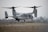 A military aircraft with tiltrotor engines is pictured hovering close to the ground on a runway, surrounded by a hazy environment and dry grassland.