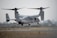 A military aircraft with tiltrotor engines is pictured hovering close to the ground on a runway, surrounded by a hazy environment and dry grassland.
