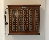 A vintage wooden telephone switchboard featuring multiple round dials with numbers and an antique telephone receiver attached to the side. The board has rows of metallic elements and numeric labels beneath each dial.