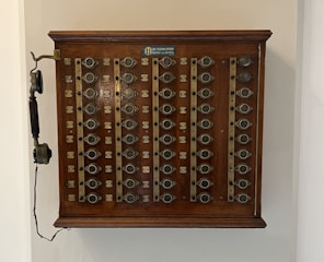 A vintage wooden telephone switchboard featuring multiple round dials with numbers and an antique telephone receiver attached to the side. The board has rows of metallic elements and numeric labels beneath each dial.