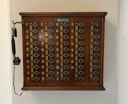 A vintage wooden telephone switchboard featuring multiple round dials with numbers and an antique telephone receiver attached to the side. The board has rows of metallic elements and numeric labels beneath each dial.