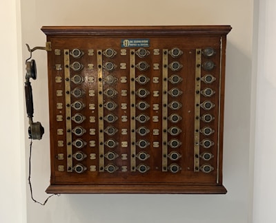 A vintage wooden telephone switchboard featuring multiple round dials with numbers and an antique telephone receiver attached to the side. The board has rows of metallic elements and numeric labels beneath each dial.