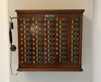 A vintage wooden telephone switchboard featuring multiple round dials with numbers and an antique telephone receiver attached to the side. The board has rows of metallic elements and numeric labels beneath each dial.