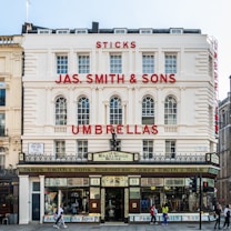 A historic building with a cream-colored facade displaying large red signage that reads 'JAS. SMITH & SONS UMBRELLAS.' The structure features arched windows and decorative architectural elements. Below, there are storefront windows exhibiting various umbrellas and sticks. People are walking by on the sidewalk in front.