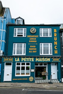 A vibrant blue building with white-framed windows featuring a café bar and bistro named La Petite Maison. The façade displays yellow text advertising breakfast, lunch, dinner, and all-day food on offer. A decorative clock is mounted above, and the street outside is paved.