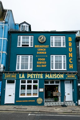 A vibrant blue building with white-framed windows featuring a café bar and bistro named La Petite Maison. The façade displays yellow text advertising breakfast, lunch, dinner, and all-day food on offer. A decorative clock is mounted above, and the street outside is paved.