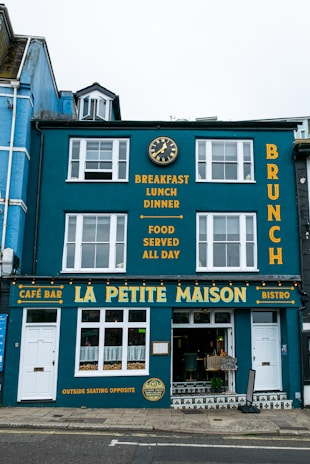 A vibrant blue building with white-framed windows featuring a café bar and bistro named La Petite Maison. The façade displays yellow text advertising breakfast, lunch, dinner, and all-day food on offer. A decorative clock is mounted above, and the street outside is paved.