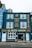 A vibrant blue building with white-framed windows featuring a café bar and bistro named La Petite Maison. The façade displays yellow text advertising breakfast, lunch, dinner, and all-day food on offer. A decorative clock is mounted above, and the street outside is paved.