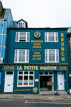 A vibrant blue building with white-framed windows featuring a caf&eacute; bar and bistro named La Petite Maison. The fa&ccedil;ade displays yellow text advertising breakfast, lunch, dinner, and all-day food on offer. A decorative clock is mounted above, and the street outside is paved.